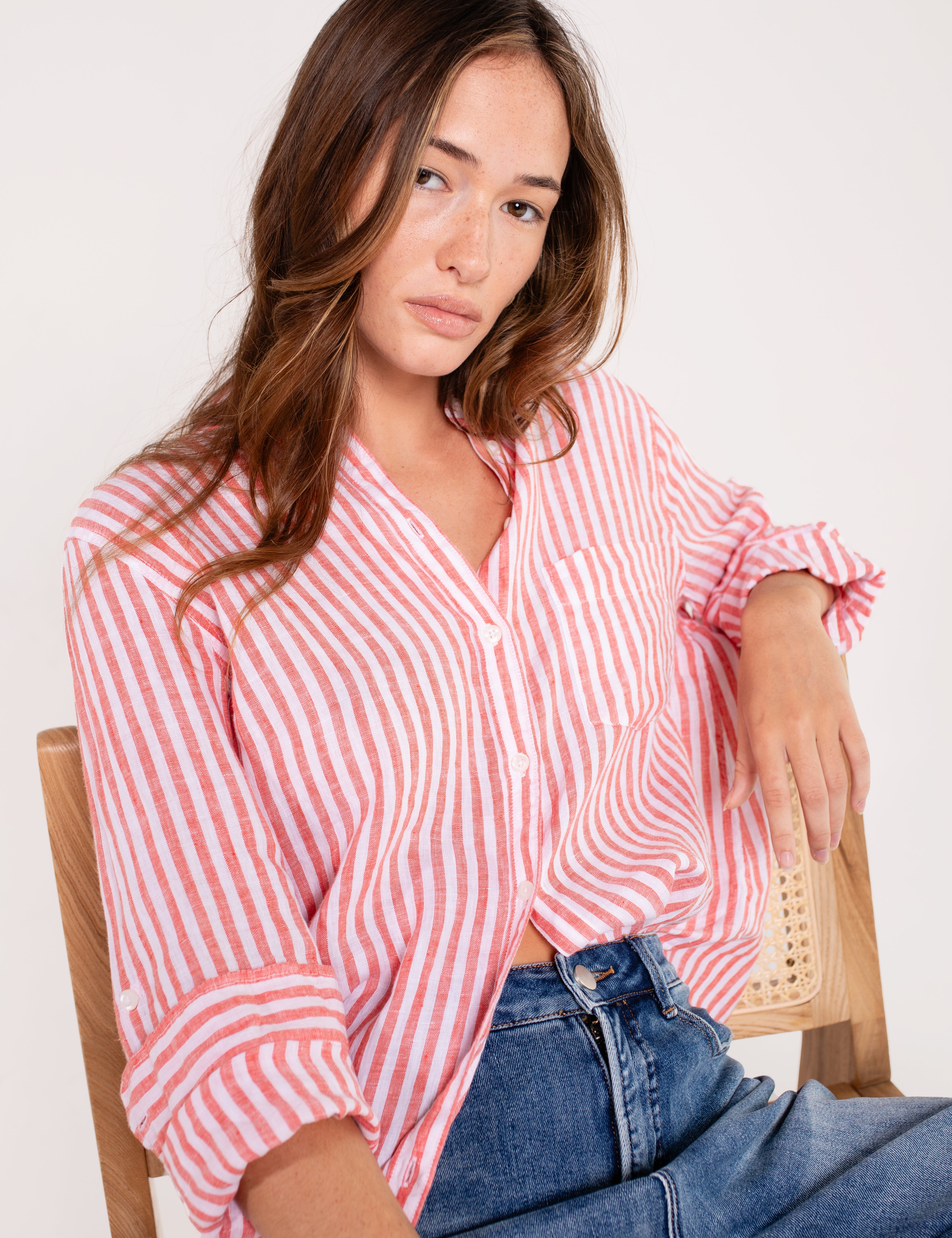 A woman sitting and wearing a Hut red and white striped boyfriend linen shirt with button-up sleeves and a scoop hem, paired with blue jeans.