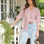 A woman standing on a porch wearing a Hut Rhubarb striped boyfriend linen shirt with long sleeves and light blue jeans. She is holding a watering can and there are plants in the background.