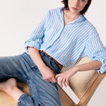 Model wearing a Hut Clothing sky blue yarn dyed stripe boyfriend linen shirt with rolled-up sleeves, paired with blue jeans, sitting on a chair.
