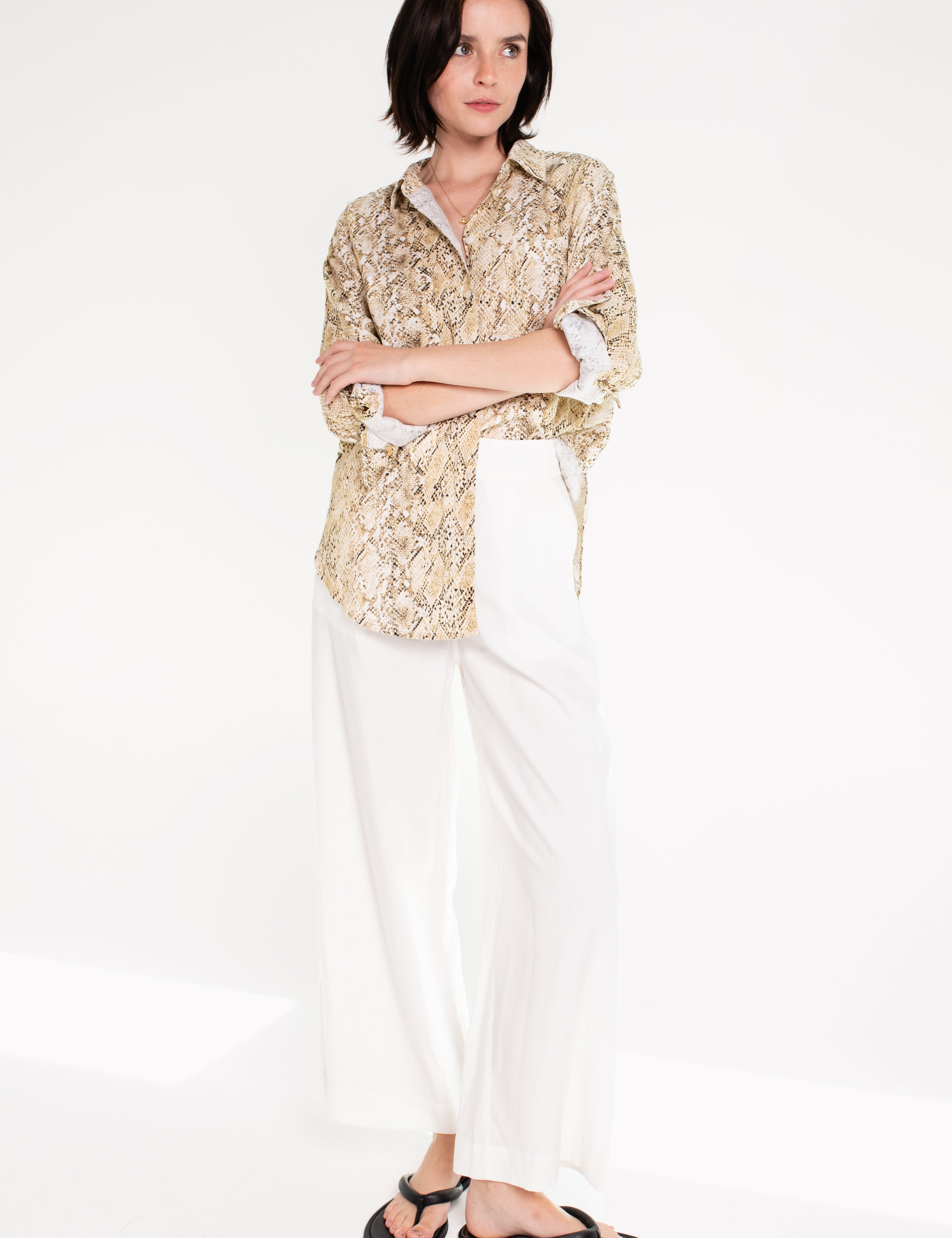 A woman wearing a Hut snake skin print boyfriend linen shirt in an oversized fit, paired with white trousers, standing against a white background.