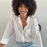 A model smiling, wearing a white linen Hut clothing shirt with one chest pocket and light blue jeans.