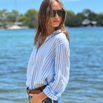 A woman standing by the water wearing a striped Hut Clothing boyfriend linen shirt in blue and white.